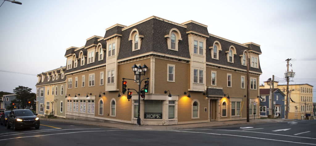 A three-story tan and cream building with a mansard roof and arched windows on a corner. A "HOPE" sign is displayed in the ground-level windows, with streetlights and traffic lights at the intersection. A few cars wait at the lights under a clear sky.