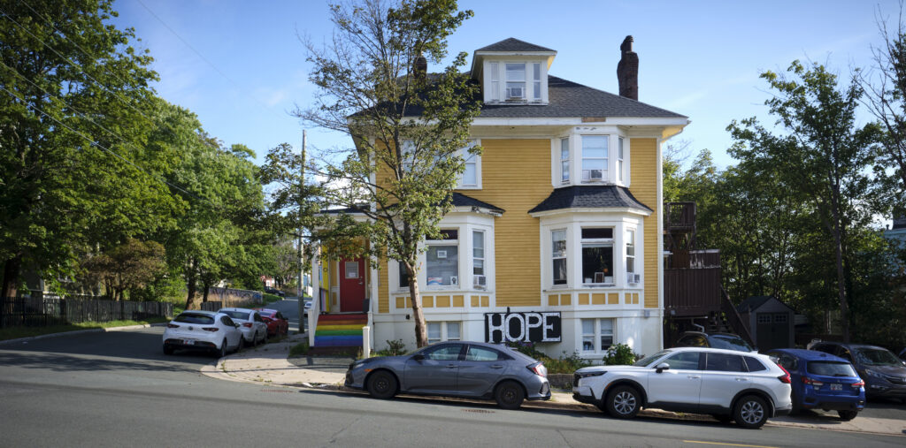 A yellow, two-story house with white trim and bay windows on a quiet street. The front porch features rainbow-painted steps leading to a red door. A "HOPE" sign is displayed near the base of the house. Several cars are parked along the street, and trees surround the area under a clear, sunny sky.